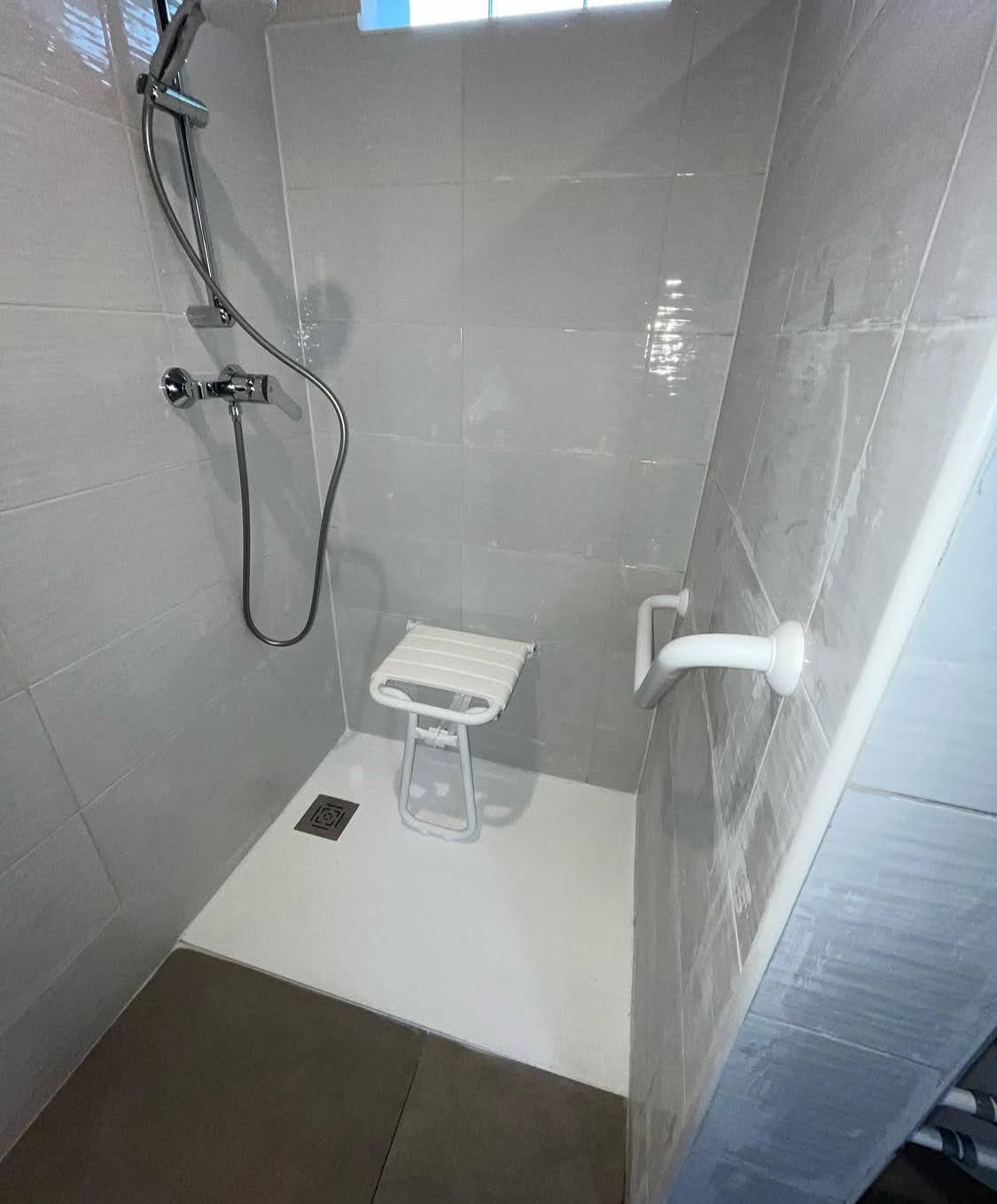 Salle de bain aux normes PMR à Brumath montrant un lavabo suspendu dégagé, un WC surélevé avec cadre d'appui et une douche à l'italienne séparée par un demi-mur.