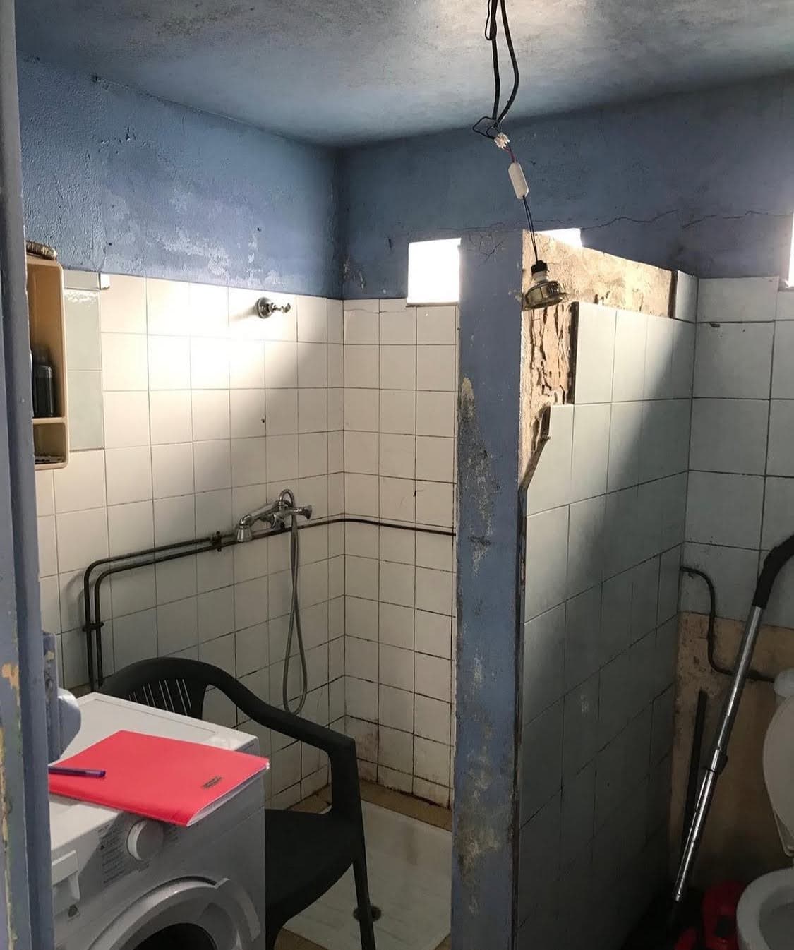 Ancienne salle d'eau insalubre avec tuyauteries apparentes, carrelage dégradé et chaise en plastique instable, nécessitant l'intervention urgente d'un installateur sanitaire.