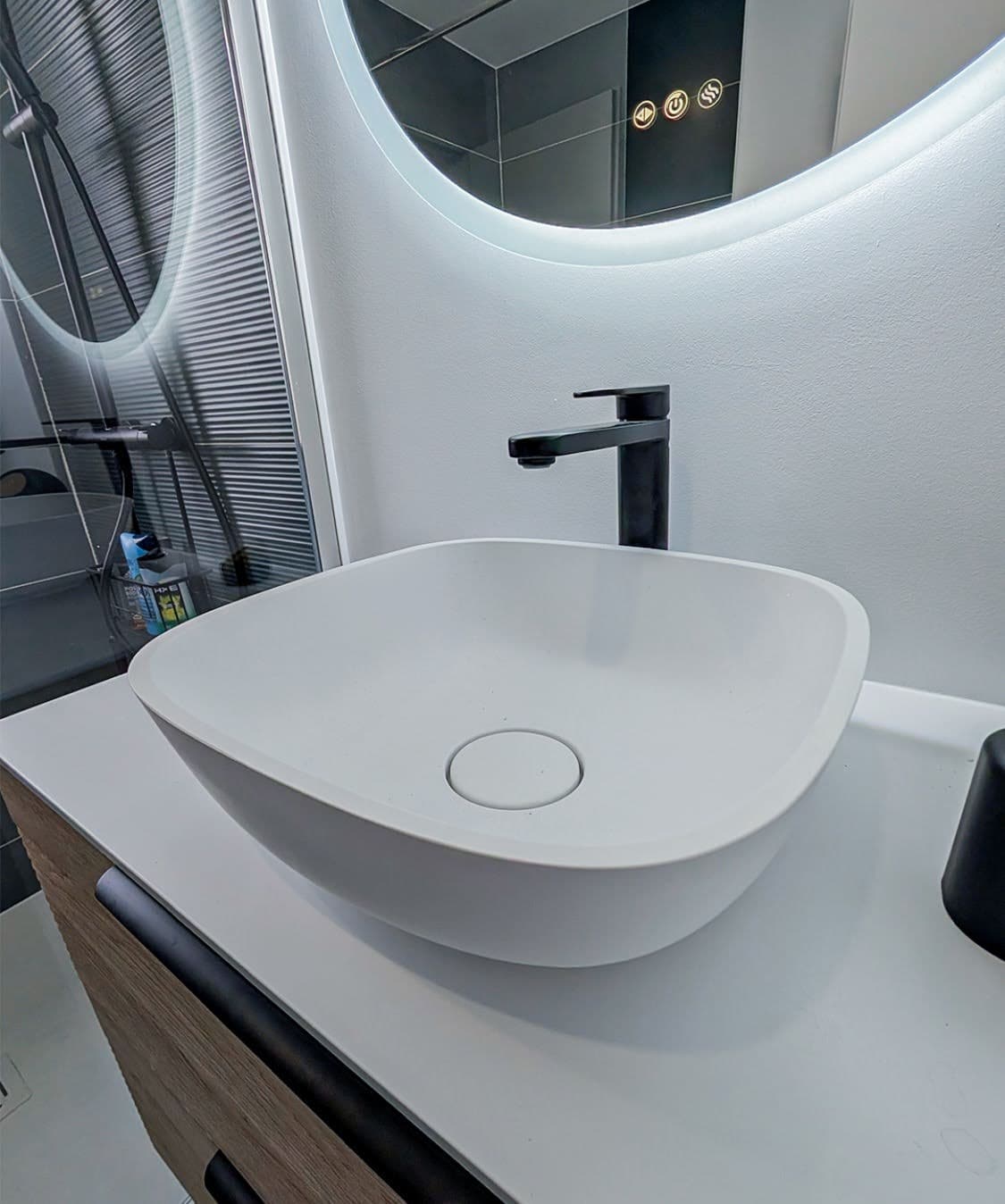 Détail de l'aménagement sur mesure avec un meuble vasque en bois nervuré, une vasque à poser blanche, un mitigeur noir et un miroir rond rétroéclairé LED.