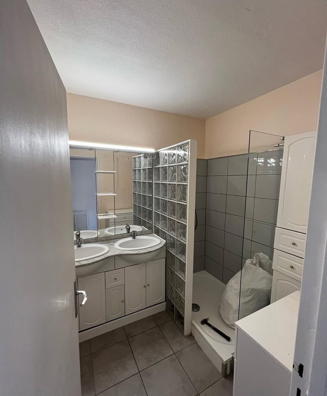 État initial d'une pièce d'eau avant une rénovation salle de bain à Sélestat, montrant un meuble double vasque démodé, un ancien sol carrelé et une paroi de douche en pavés de verre.
